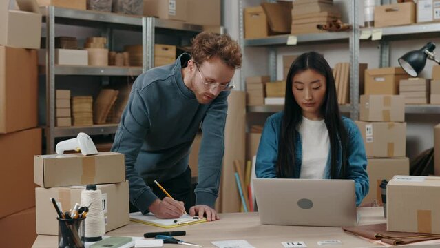 Ginger caucasian man looking at the laptop screen and writing some details at the paper while his female asian colleague working with him at the warehouse. Small business concept.