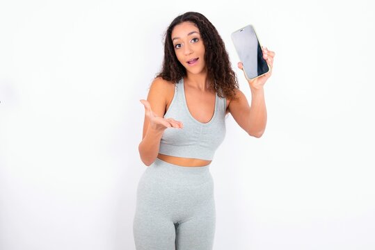 Beautiful teen girl with curly hair wearing green sport set over white background with a mobile. presenting smartphone. Advertisement concept.
