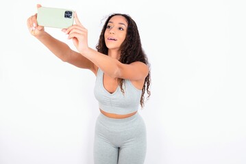 Beautiful teen girl with curly hair wearing green sport set over white background taking a selfie to post it on social media or having a video call with friends.