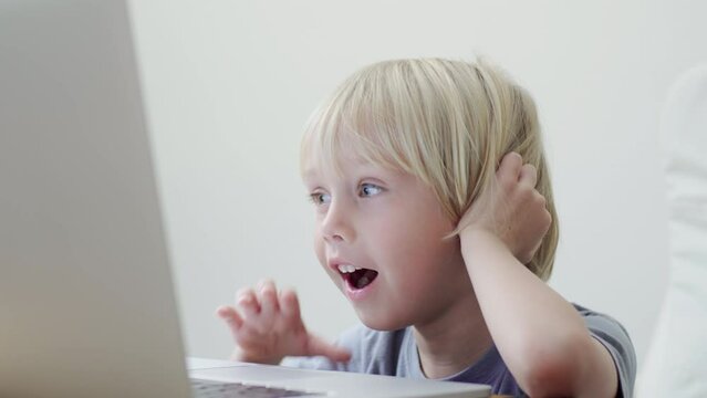 Adorable 5 years caucasian boy is watching something online at laptop at home. White background. Home education, online lessons, intertainment