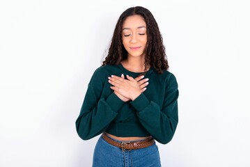 teen girl wearing knitted green sweater over white closes eyes and keeps hands on chest near heart,...