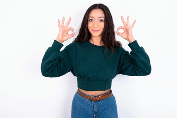 Glad teen girl wearing knitted green sweater over white background shows ok sign with both hands as expresses approval, has cheerful expression, being optimistic.