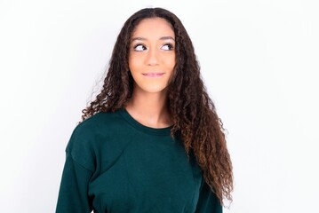 Fototapeta premium teen girl wearing knitted green sweater over white background with thoughtful expression, looks away keeps hands down bitting his lip thinks about something pleasant.