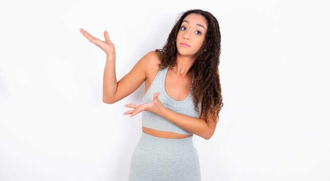 teen girl with curly hair wearing white sport set over gray background pointing aside with both hands showing something strange and saying: I don't know what is this. Advertisement concept.