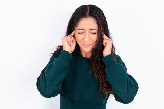 Stop making this annoying sound! Unhappy stressed out teen girl wearing knitted green sweater over white background making worry face, plugging ears with fingers, irritated with loud noise.