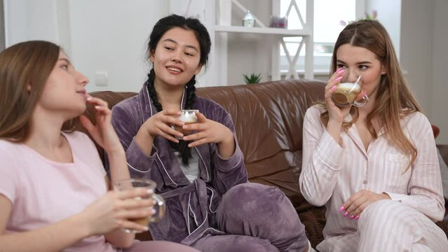 Joyful Asian and Caucasian relaxed women toasting with coffee talking sitting on couch indoors. Happy carefree female friends in pajamas enjoying leisure gossiping sharing rumors