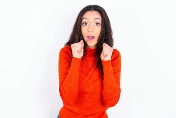 Portrait of desperate and shocked teen girl wearing knitted red sweater over white background looking panic, holding hands near face, with mouth wide open.