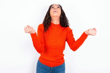 teen girl wearing knitted red sweater over white background looks with excitement up, keeps hands raised, notices something unexpected.