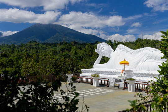 White sleeping buddha statue in a reclining pose with a mountain and trees in the background, Vihara Dharma Giri Temple; Tabanan, Bali, Indonesia