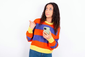 beautiful teen girl wearing knitted colourful sweater over white background points thumb away and shows blank space aside, holds mobile phone for sending text messages.