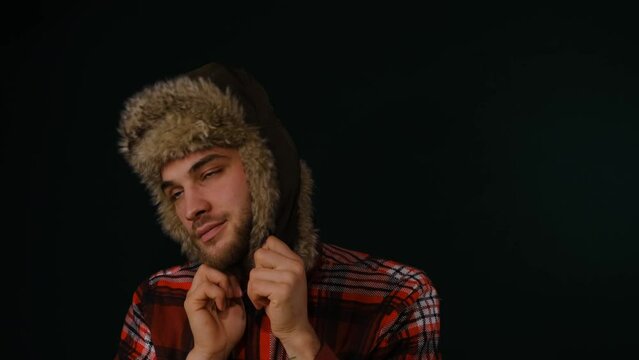 Young Caucasian bearded man wears fluffy fur hat with earflaps on head and dances on dark studio background. Classic Russian stereotype simple Siberian guy in national clothes of Northern peoples.