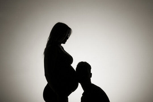 A silhouette in a studio of a couple who are having a baby and the father is listening to the mother's belly who is well along in her pregnancy; Edmonton, Alberta, Canada