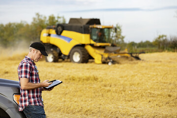 A farmer making a call and using his tablet to help manage the wheat harvest while a combine is working in the background: Alcomdale, Alberta, Canada
