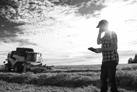 A farmer making a call and using his tablet to help manage the wheat harvest while a combine is working in the background: Alcomdale, Alberta, Canada