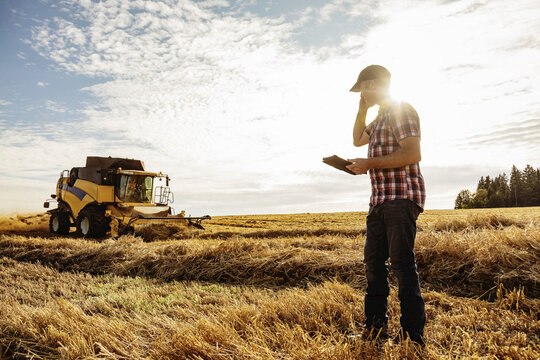 A farmer making a call and using his tablet to help manage the wheat harvest while a combine is working in the background: Alcomdale, Alberta, Canada
