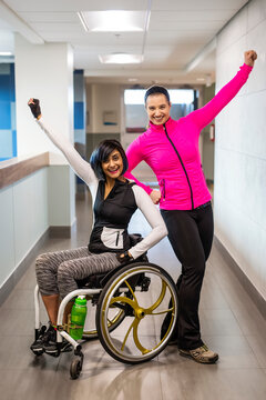 A paraplegic woman and her trainer do a celebratory pose for the camera while in a hallway in a recreational facility: Sherwood Park, Alberta, Canada