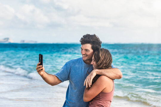 A young Millennial couple taking a selfie with a smart phone on Lanakai Beach with the Moku Manu Islands in the background; Lanakai, Oahu, Hawaii, USA