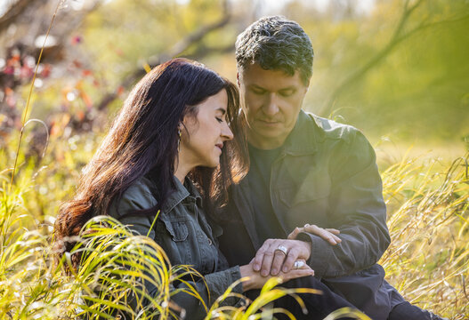 Mature couple sitting in the tall grass holding hands and praying together while on a walk in the woods in autumn; St Albert, Alberta, Canada