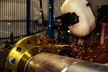 A welder wearing mask and eye protection is grinding a flange butt weld in a fabrication plant; Innisfail, Alberta, Canada