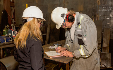 A female plant manager provides administrative documents for signing by an employee for job tracking in a fabrication plant; Innisfail, Alberta, Canada