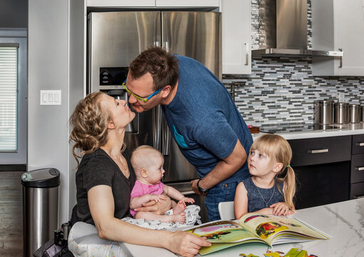 A husband kissing his paraglegic wife while they are reading and making a puzzle with their baby and young daughter; Edmonton, Alberta, Canada