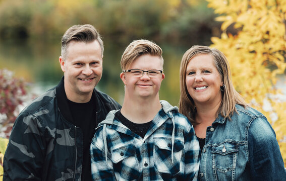A young man with Down Syndrome posing for a family portrait with his father and mother while enjoying each other's company in a city park on a warm fall evening; Edmonton, Alberta, Canada