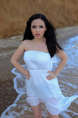 Fototapeta na wymiar a beautiful young fashionable girl with black hair in a white dress with big breasts stands on the beach in summer by the water of the sea ocean