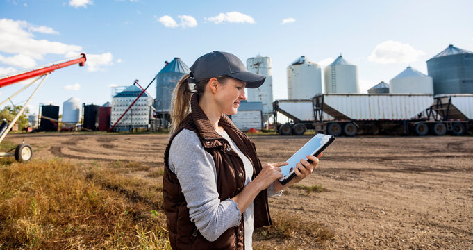 Mature woman farmer working on a tablet computer and standing in front of grain bins and loading facility at her grainery; Alcomdale, Alberta, Canada