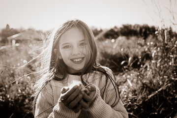 Young girl standing in a farm field holding fluffy vegetation in her hand, smiling and looking at the camera; Alcomdale, Alberta, Canada