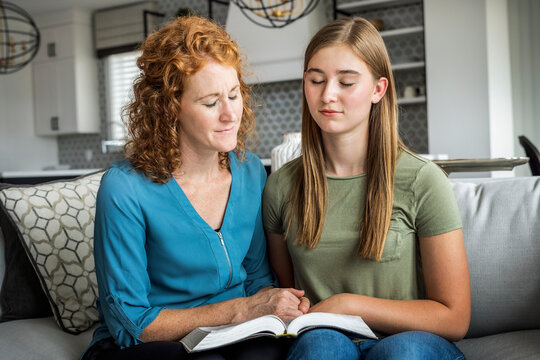 Mother and teenage daughter sitting on a couch at home reading the Bible and praying together; Edmonton, Alberta, Canada