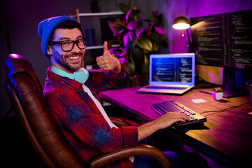 Photo of confident smiling coder dressed hat eyewear showing thumb up working modern gadget indoors...