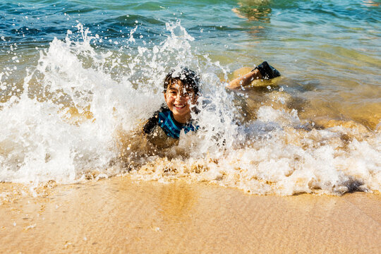 A boy looks up at the camera with a smile as he lays on the shore and a large wave crashes over him at D. T. Fleming Beach; Kapalua, Maui, Hawaii, United States of America