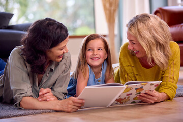Same Sex Family With Two Mature Mums And Daughter Lying On Floor Reading Book At Home Together