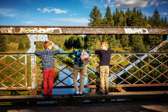 Three boys look out from a park bridge to a creek below, one boy in the middle bending over backwards to look at the camera in a silly pose; Edmonton, Alberta, Canada