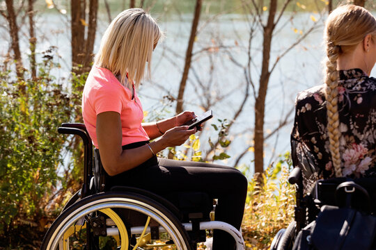 Paraplegic young woman using her smart phone while friends enjoy a beautiful fall day outdoors at a city park; Edmonton, Alberta, Canada