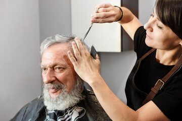 Working with hair scissors. Professional hairstyler woman cutting hair of mature man. He is getting...