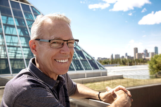 Outdoor portrait of senior man with eyeglasses, city skyline and modern architecture in the background; Edmonton, Alberta, Canada