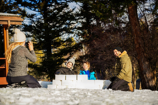 A Mother Takes A Picture Of A Father And Two Sons Building An Igloo Together Out Of Snow Blocks At A Mountain Resort; Fairmont Hot Springs, British Columbia, Canada