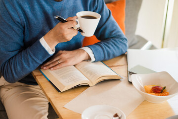 Hands of an unrecognizable person studying with a book and holding a coffee.