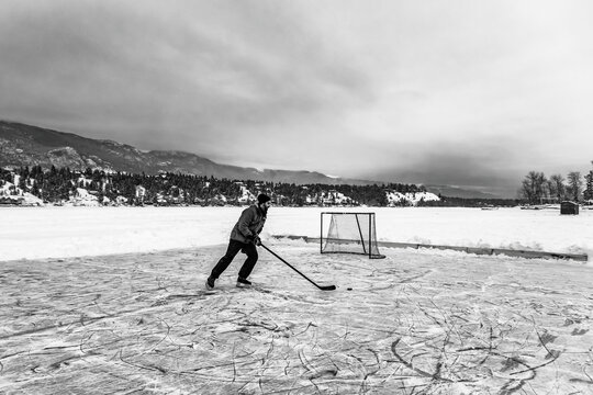 Black and white image of a man playing hockey alone on a frozen Windermere Lake during the winter, Windermere Lake Provincial Park; Invermere, British Columbia, Canada