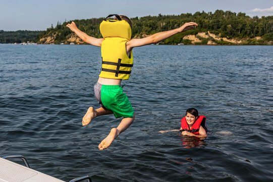 Young boy jumping off a boat and swimming in a lake with a lifejacket and his brother watching him, Lac Ste. Anne; Alberta Beach, Alberta, Canada