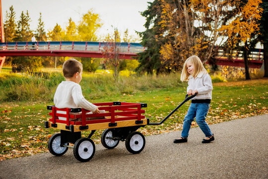 Young girl pulling her brother in a wagon at a city park along a river during the fall season; St. Albert, Alberta, Canada