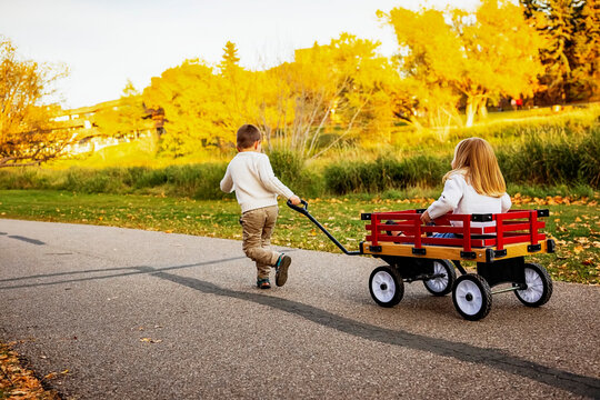 Young boy pulling his sister in a wagon at a city park along a river during the fall season; St. Albert, Alberta, Canada