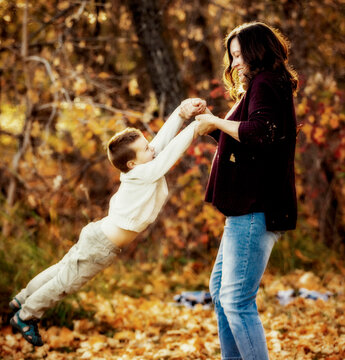 A mother spending quality time and playing with her young son and swinging him around during a family outing outdoors in a city park during the fall season; Edmonton, Alberta, Canada