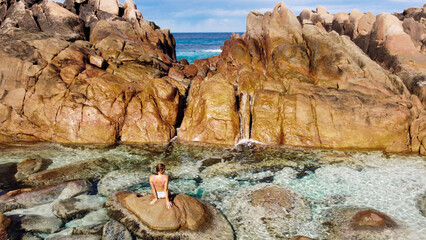 Woman in bikini sitting on large rock in clear torquiest rock pools of Injidup Natural Spa in Yallingup Western Australia. Tan rocks and blue skies and ocean in background. 