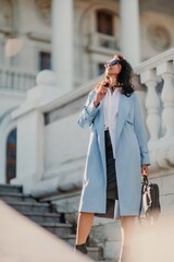 Portrait of a brunette business lady with glasses. Woman walks around the city, lifestyle. She wears a blue cloak, a black leather skirt, and black heeled boots.