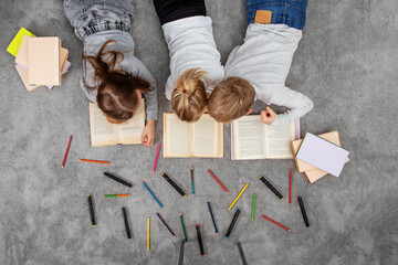 Group of children are reading books and drawing with pencils while lying on floor. Top view