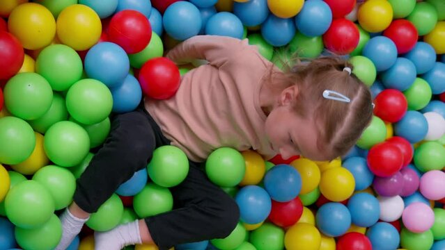 Cute little girl playing on multi coloured plastic balls in big dry paddling pool in playing centre. Having fun in playroom