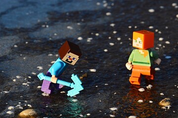 Fototapeta premium LEGO Minecraft figures of Steve and Alex on thick layer of real ice Steve is trying to break ice with his diamond pickaxe.
