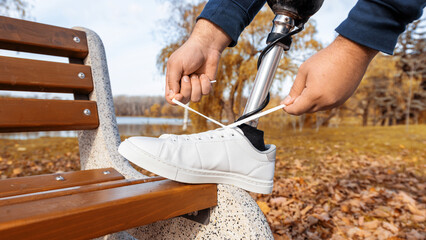 View of a man with prosthetic legs and white sneakers. Tying shoelaces with his foot on the bench in a park. Fallen yellow leaves around